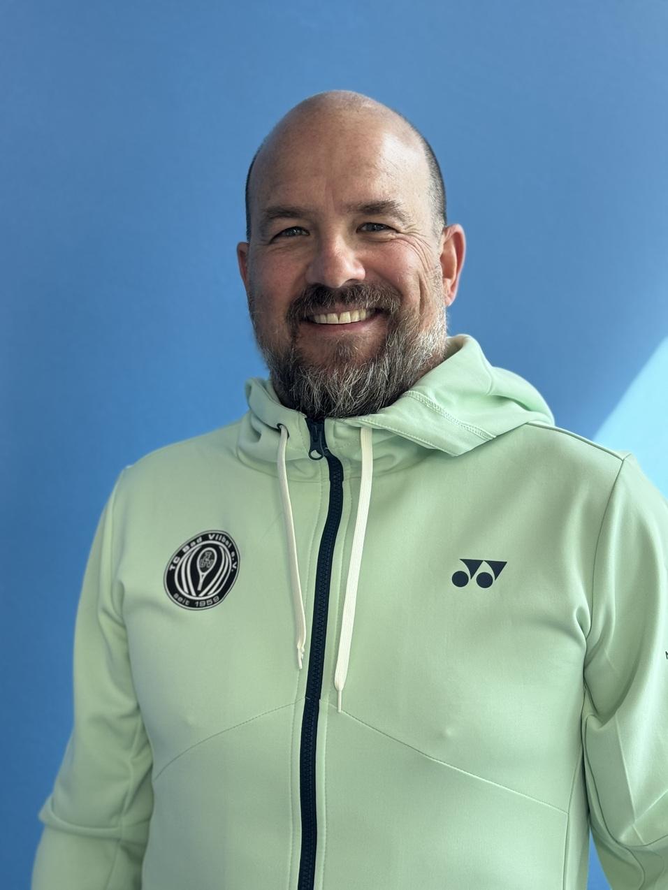 Mann mit Bart lächelt in mintgrünem Kapuzenpullover vor blauem Hintergrund.