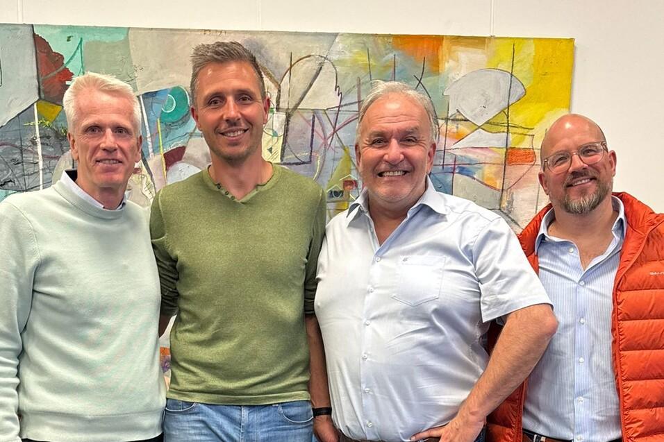 Vier M&auml;nner stehen nebeneinander in einem modernen Raum vor einem bunten Kunstwerk. Alle l&auml;cheln in die Kamera.