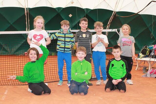 Gruppe von acht Kindern in Sportkleidung auf einem Tennisplatz mit Netz im Hintergrund.