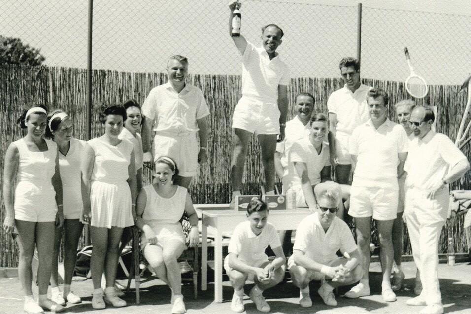 Gruppenschnappschuss von zw&ouml;lf Personen in wei&szlig;en Sportoutfits, ein Sieger mit Pokal steht auf einem Tisch.