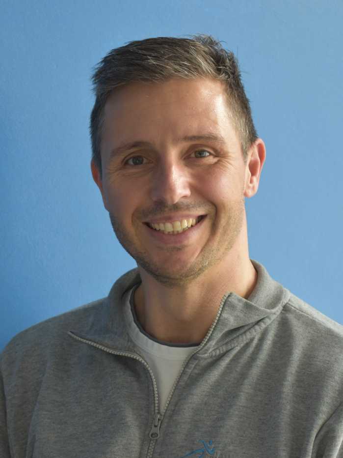 L&auml;chelnder Mann mit kurzen Haaren, tr&auml;gt einen grauen Pullover vor einer blauen Wand.