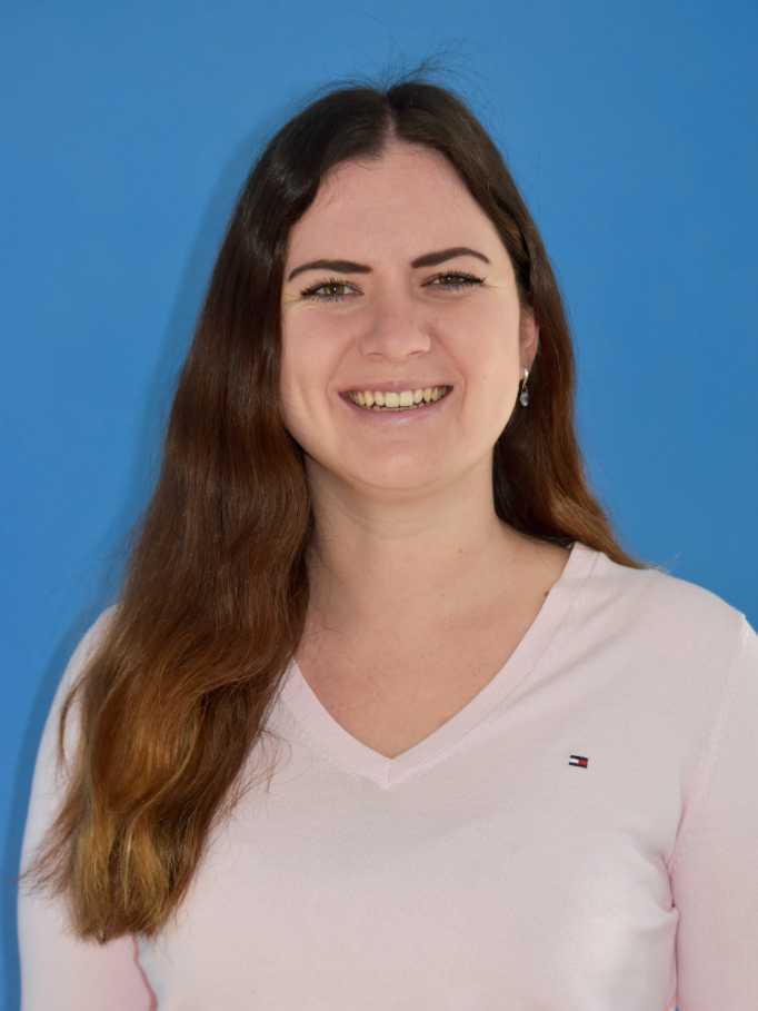 L&auml;chelnde Frau mit langen, braunen Haaren in einem rosa Pullover vor einem blauen Hintergrund.