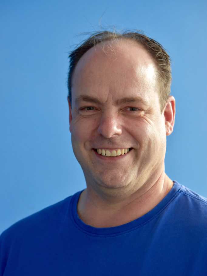 Lächelnder Mann mit kurzer Haarfrisur trägt ein blaues T-Shirt vor einem hellblauen Hintergrund.
