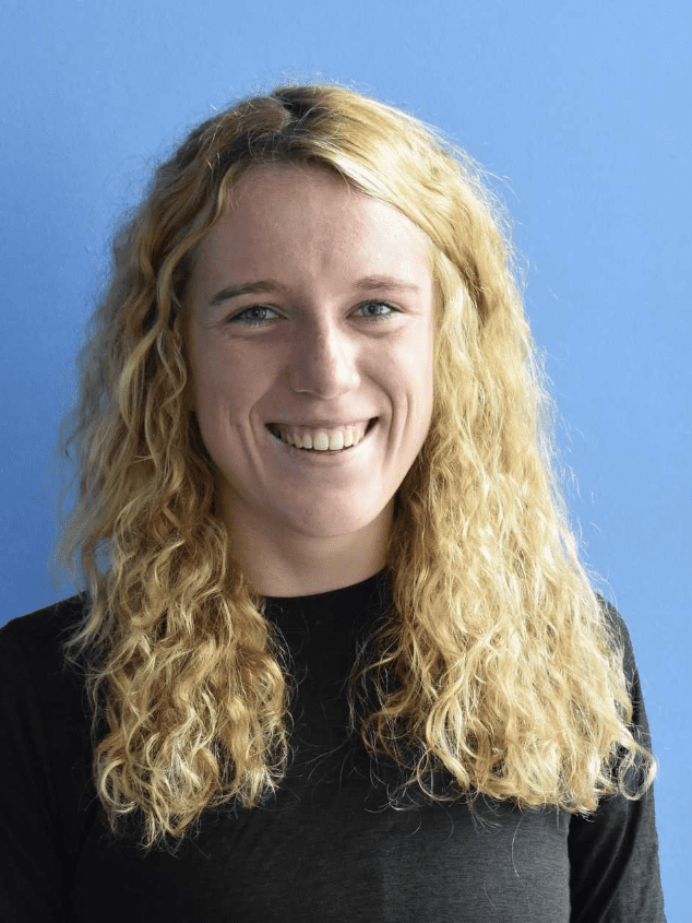 L&auml;chelnde Frau mit lockigem, blondem Haar vor blauem Hintergrund. Sie tr&auml;gt ein schwarzes Oberteil.