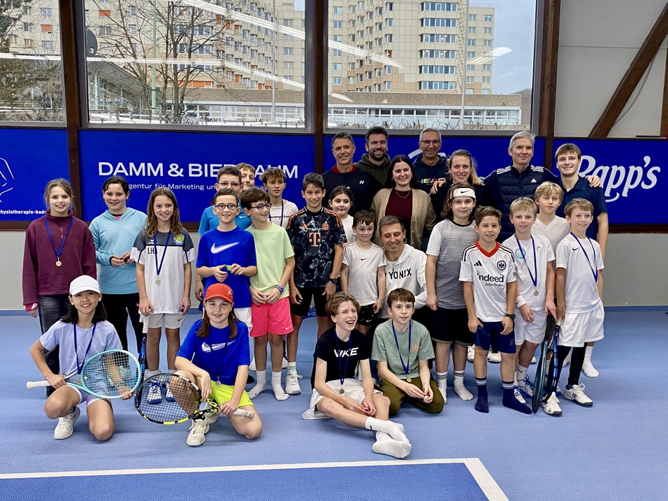 Gruppe von Jugendlichen und Erwachsenen l&auml;chelt auf einem Tennisplatz, umgeben von Sportausr&uuml;stung und Fenstern.