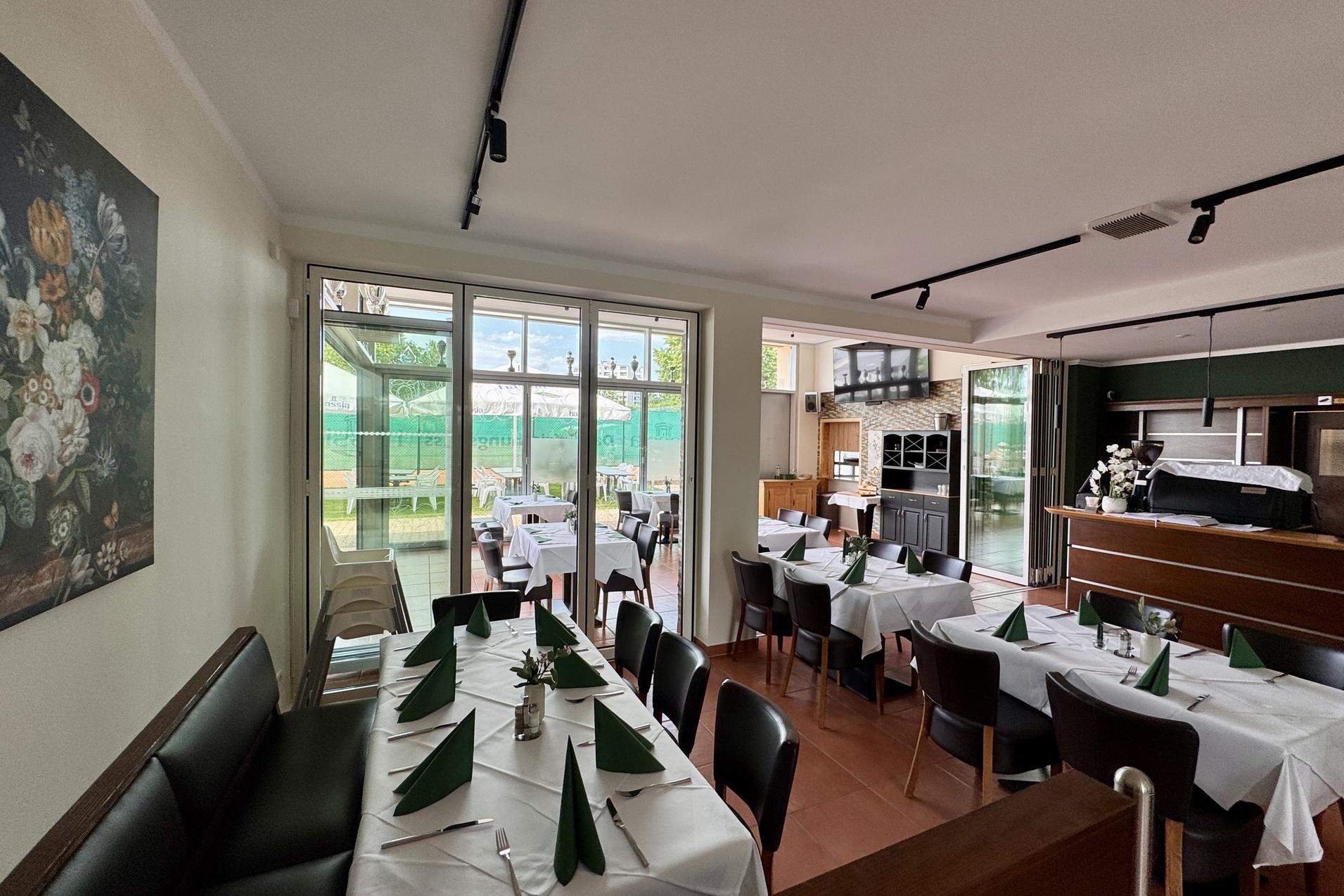 Gem&uuml;tlicher Restaurantbereich mit Tischen, gedeckt mit wei&szlig;en Tischdecken und gr&uuml;nen Servietten, daneben Fenster mit Ausblick.