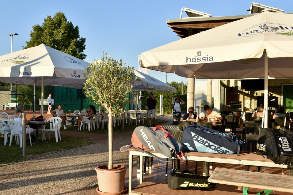 Terrasse eines Tennisplatzes mit Sonnenschirmen, Tischen, St&uuml;hlen und Tennistaschen im Vordergrund.