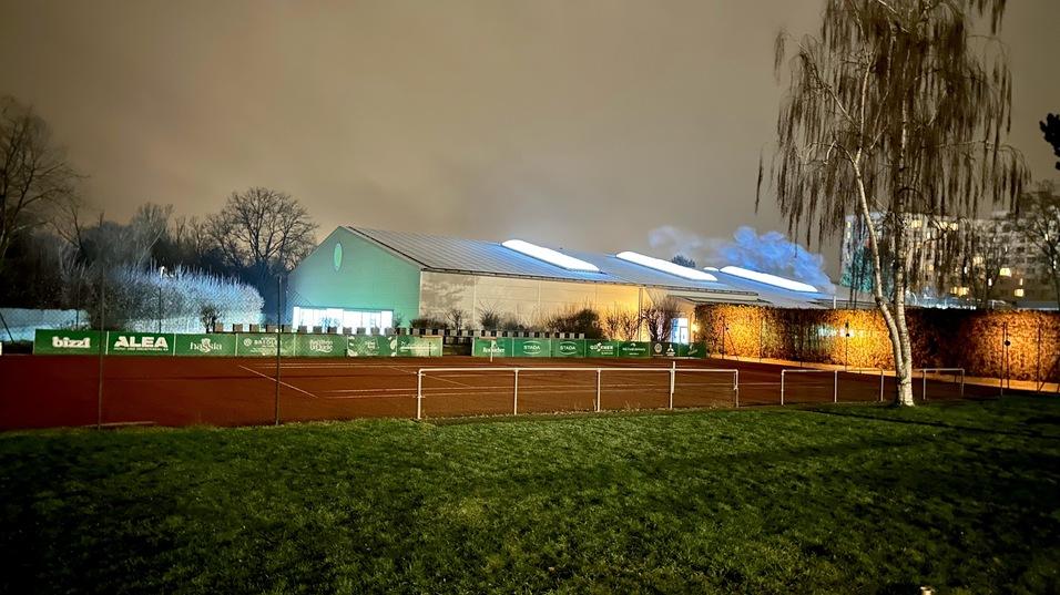 Beleuchteter Tennisplatz mit grüner Umzäunung und einer angrenzenden Sporthalle bei nächtlicher Stimmung.