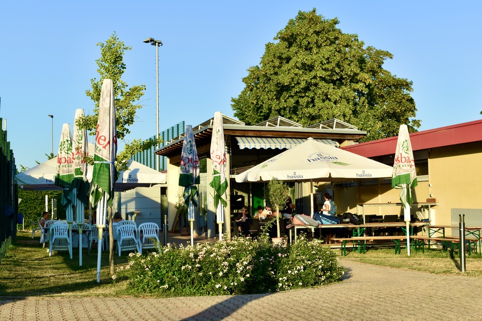 Gartenbereich eines Restaurants mit wei&szlig;en Tischen, St&uuml;hlen und Sonnenschirmen, umgeben von B&auml;umen und Pflanzen.