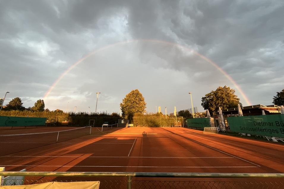 Regenbogen über einem Tennisplatz mit roten Sandboden unter bewölktem Himmel. Bäume im Hintergrund.