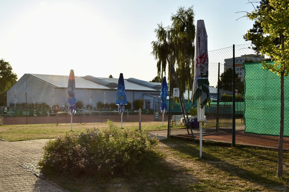 Tennisanlage mit mehreren Pl&auml;tzen, Sonnenschirmen und einer modernen Sporthalle im Hintergrund. Sonnenuntergangsstimmung.