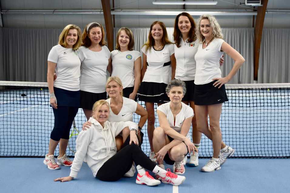 Eine Gruppe von neun Frauen in Sportkleidung steht und sitzt vor einem Tennisnetz in einer Halle.