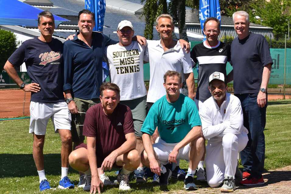 Gruppenfoto von neun M&auml;nnern in sportlichen Outfits auf einer Tennisanlage im Freien, umgeben von gr&uuml;ner Wiese und Tennispl&auml;tzen.