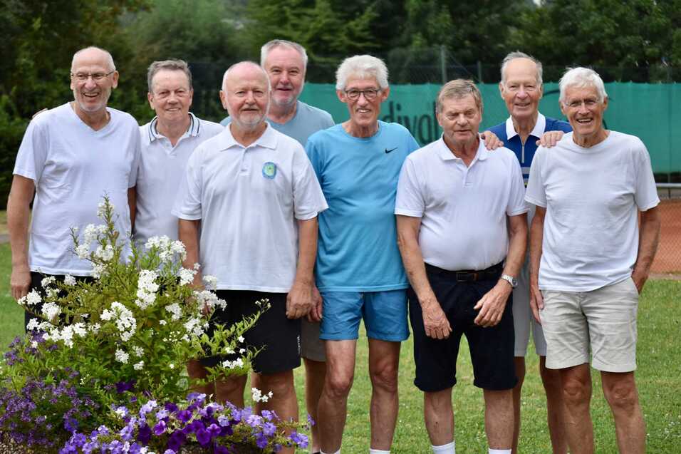 Gruppe von M&auml;nnern in Sportkleidung auf einem Tennisplatz, umgeben von bunten Blumen.