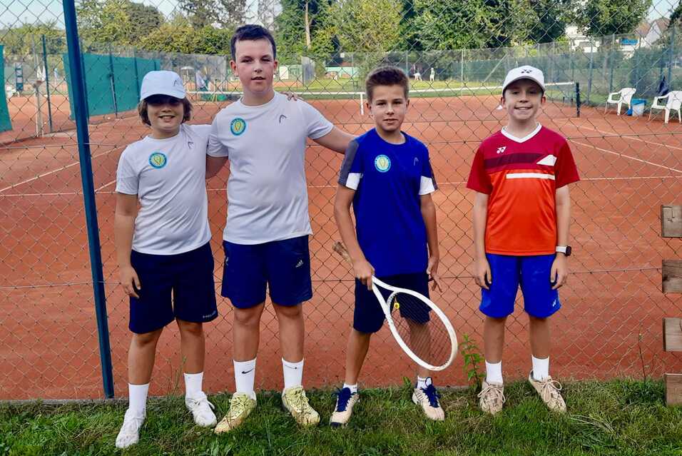 Vier Kinder in Tennisbekleidung stehen grinsend vor einem Tennisplatz mit rotem Bodenbelag und Zaun im Hintergrund.