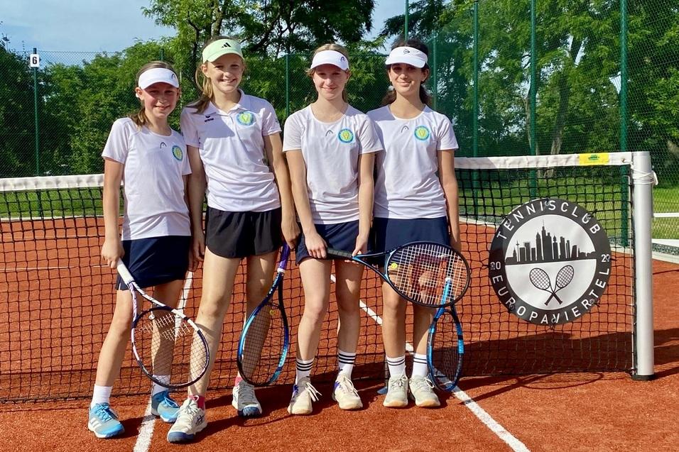 Vier M&auml;dchen stehen mit Tennisschl&auml;gern an einem Tennisnetz auf einem roten Platz, das Logo des Tennisclubs ist sichtbar.