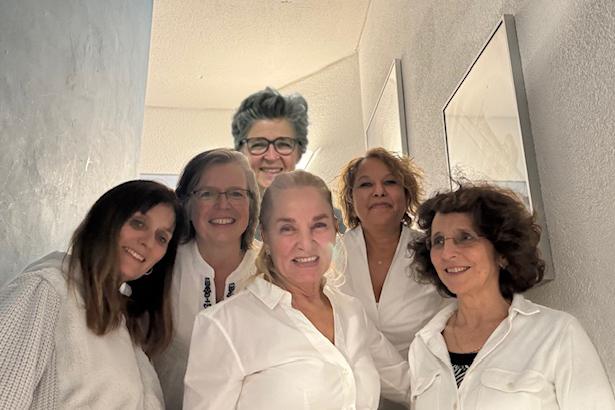 Sechs Frauen in wei&szlig;en Blusen stehen in einem hellen Flur und l&auml;cheln in die Kamera.