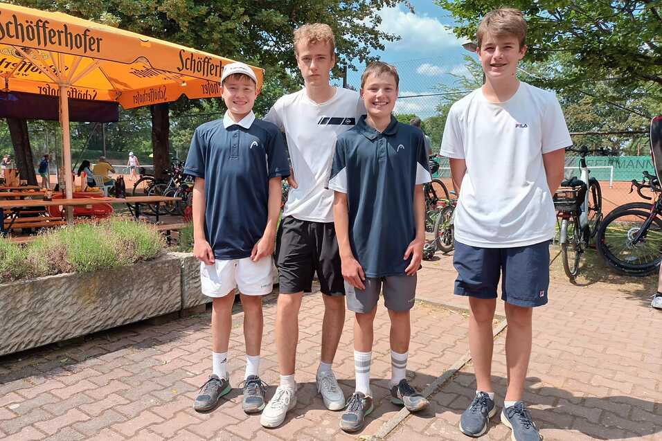 Vier junge Tennis-Spieler stehen l&auml;chelnd auf einem Platz, im Hintergrund B&auml;nke und ein Sonnenschirm sichtbar.