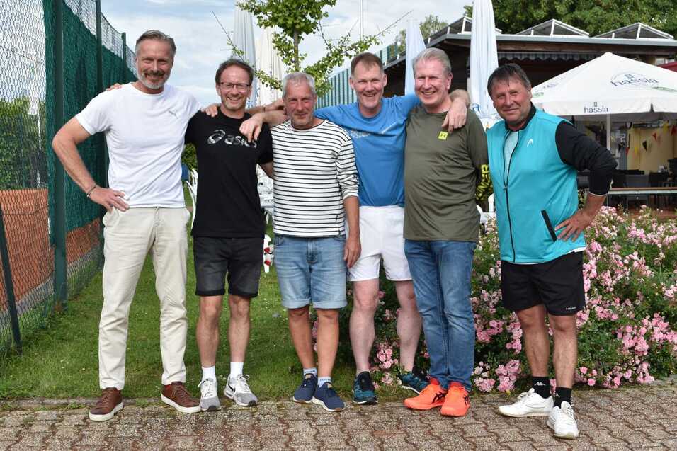 Sechs M&auml;nner posieren l&auml;chelnd vor einer Tennisanlage mit Blumen und B&auml;umen im Hintergrund.