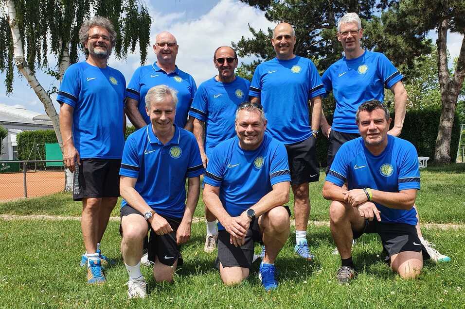 Gruppe von acht M&auml;nnern in blauen Sporttrikots steht auf einer Wiese, umgeben von B&auml;umen und einem Tennisplatz.