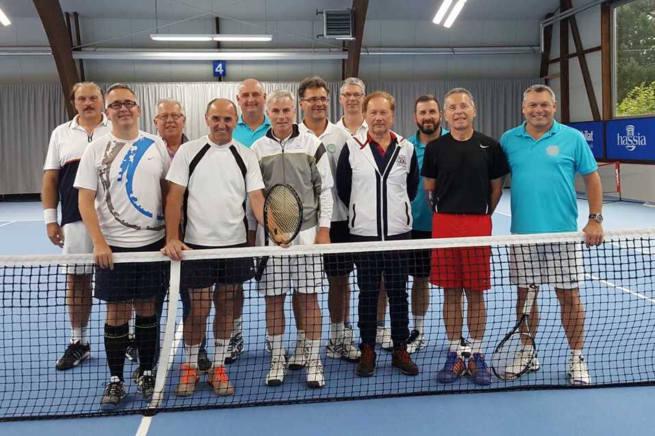 Gruppenfoto von zehn Männern in Tenniskleidung, die vor einem Tennisnetz in einer Halle stehen.