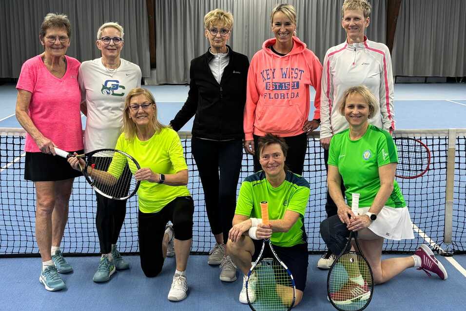 Gruppe von acht Tennis-Spielerinnen in sportlicher Kleidung, posierend vor einem Tennisnetz in einer Halle.