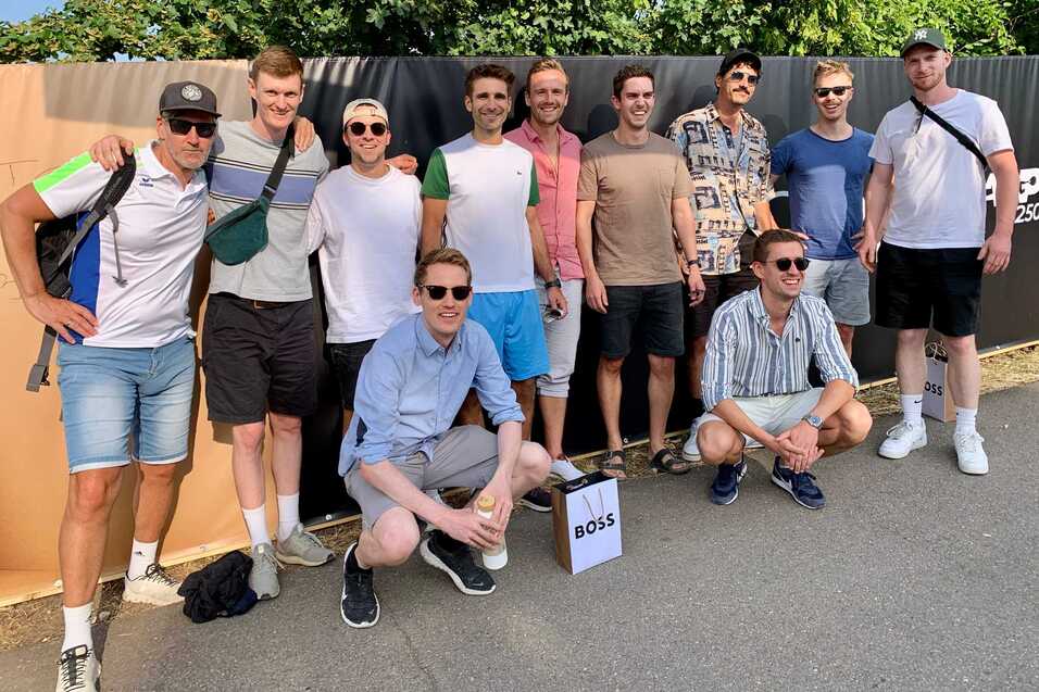 Gruppe von zehn M&auml;nnern in sportlicher Kleidung posiert l&auml;chelnd vor einer Wand, auf der "BOSS" steht.