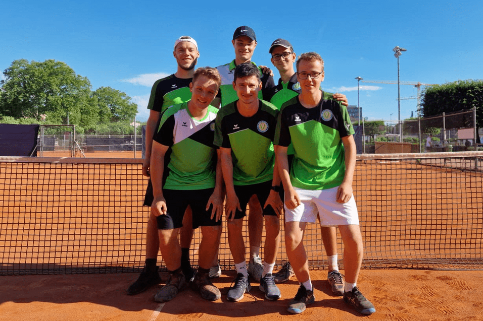 Sechs Tennisspieler in gr&uuml;nen Trikots posieren l&auml;chelnd vor einem Tennisplatz mit rotem Sand, strahlend blauer Himmel im Hintergrund.