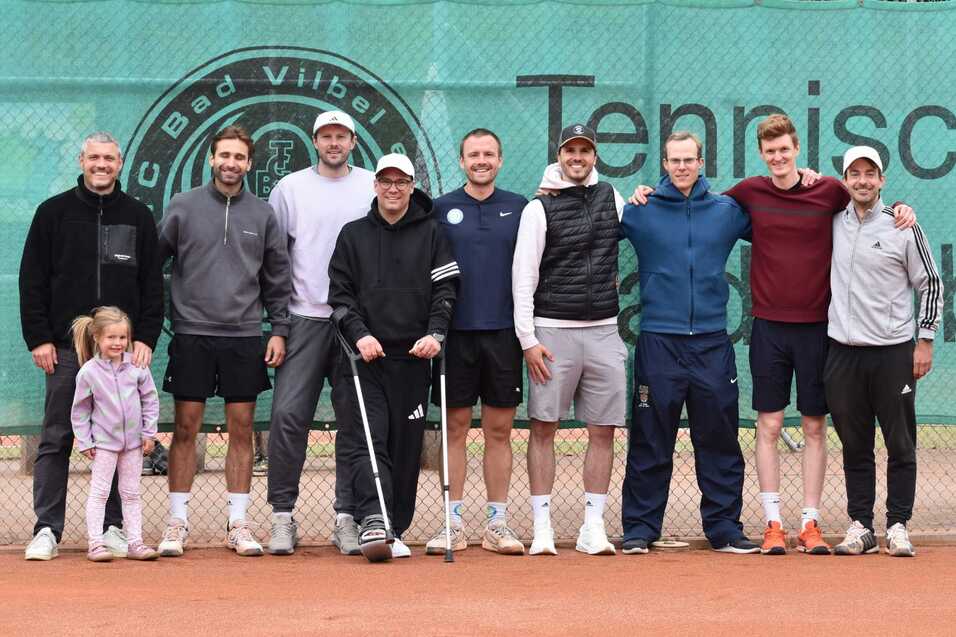 Gruppe von neun Personen, darunter ein M&auml;dchen, auf einem Tennisplatz vor einer gr&uuml;nen Wand mit Vereinslogo.