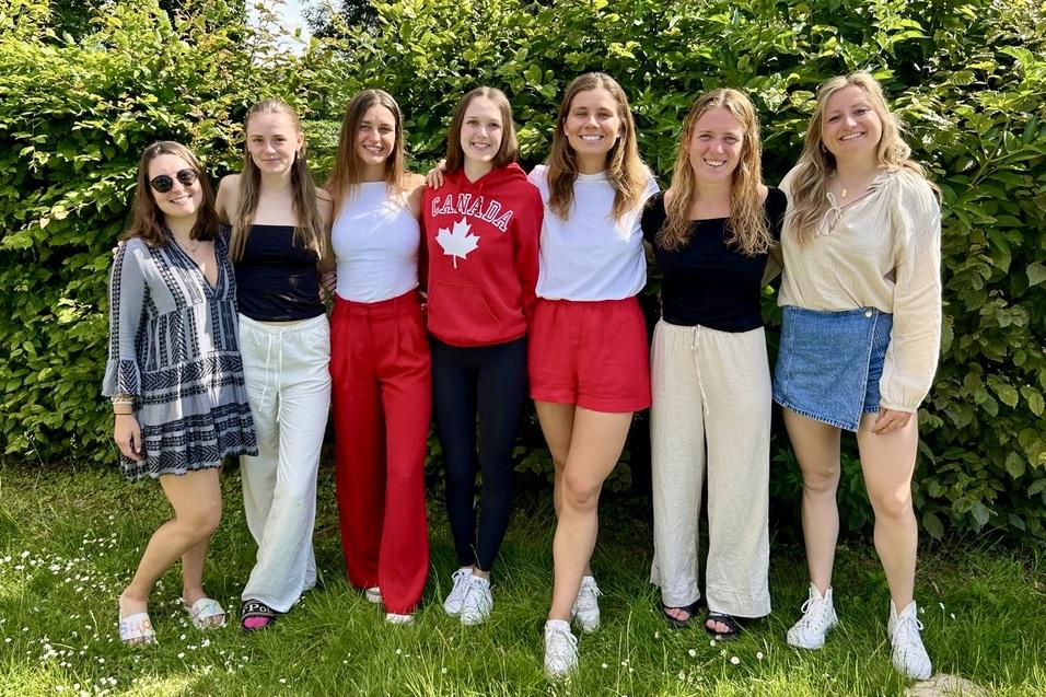 Gruppe von sieben Frauen in verschiedenen Outfits steht l&auml;chelnd vor gr&uuml;ner Hecke auf einer Wiese.