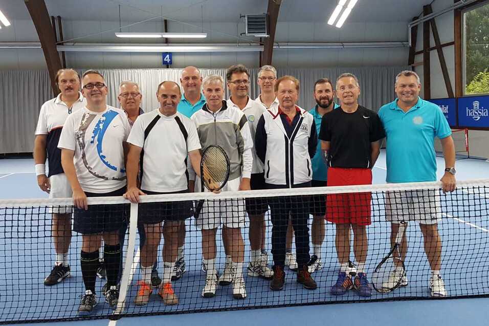 Gruppenfoto von zehn M&auml;nnern in Tenniskleidung, die vor einem Tennisnetz in einer Halle stehen.