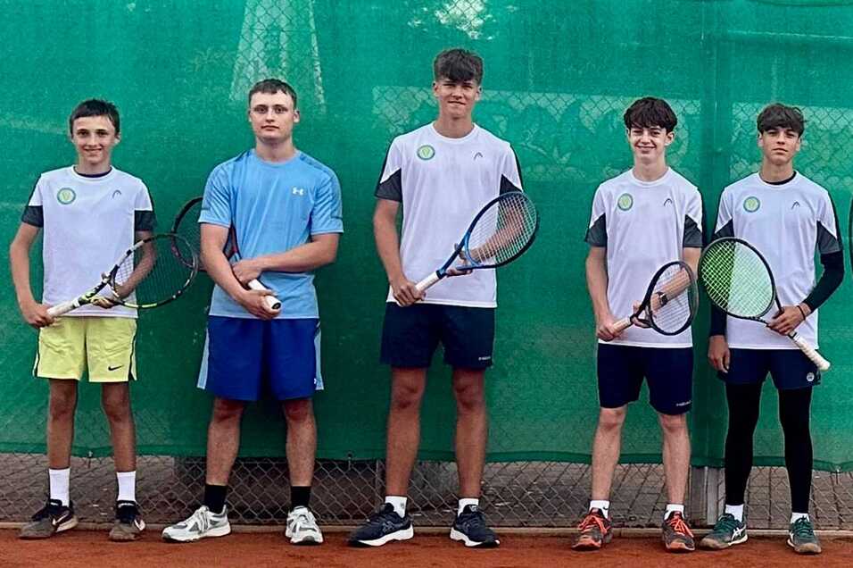 F&uuml;nf Jugendliche in Tenniskleidung stehen mit Schl&auml;gern vor einem gr&uuml;nen Sichtschutz auf einem Sandplatz.