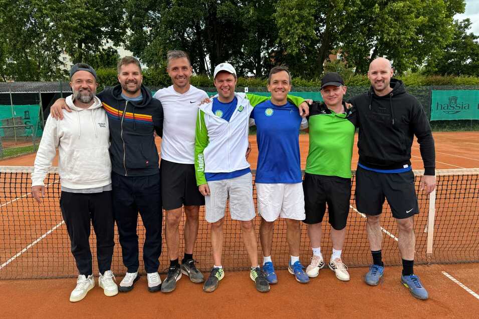 Gruppenfoto von sieben M&auml;nnern in Sportkleidung, stehend vor einem Tennisplatz mit Netz und B&auml;umen im Hintergrund.