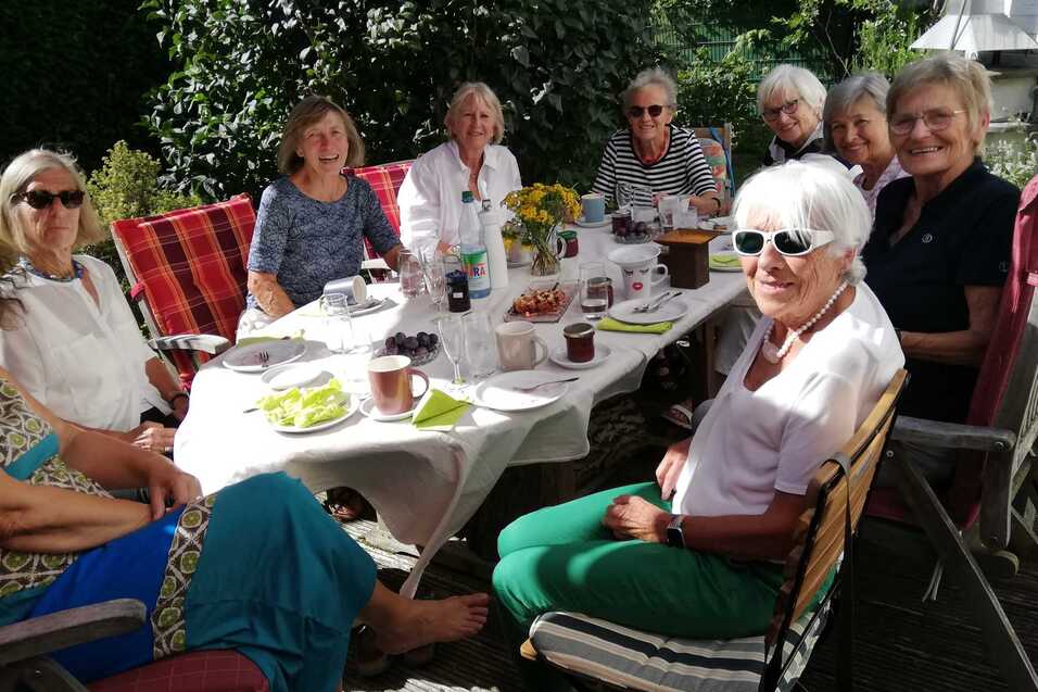 Gruppe von neun Frauen sitzt an einem gedeckten Tisch im Freien, umgeben von Gr&uuml;n, w&auml;hrend sie Kaffee und Snacks genie&szlig;en.