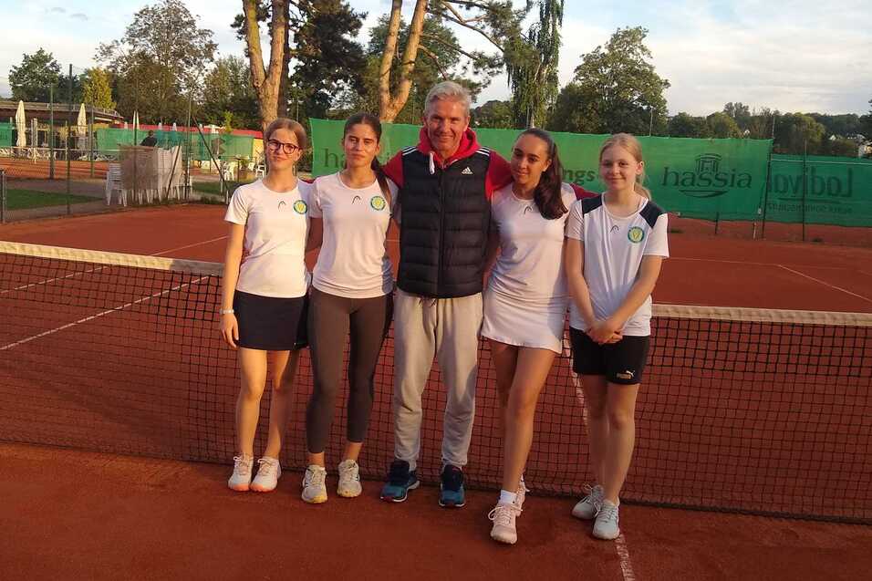 Gruppe von vier Tennisspielerinnen und einem Trainer vor einem Tennisplatz mit roten Sandbelag und gr&uuml;ner Umz&auml;unung.