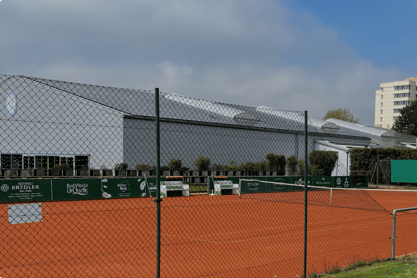 Panoramablick auf einen Tennisplatz mit rotem Sand, umgeben von einem Zaun und einem modernen Geb&auml;ude im Hintergrund.