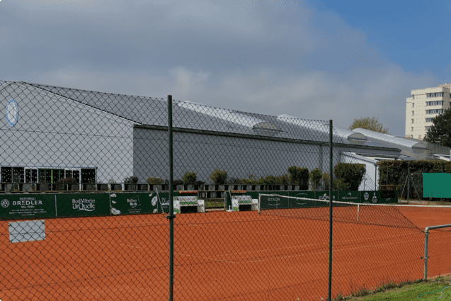 Panoramablick auf einen Tennisplatz mit rotem Sand, umgeben von einem Zaun und einem modernen Geb&auml;ude im Hintergrund.