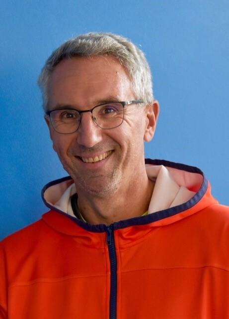 Lächelnder Mann mit Brille in orangefarbener Jacke vor blauem Hintergrund.