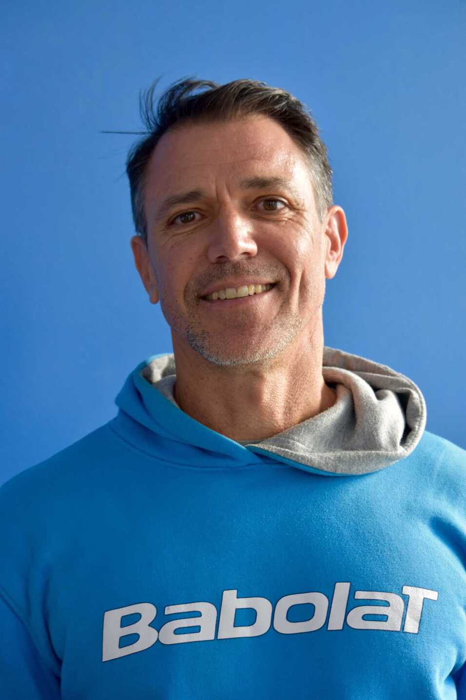 Mann mit kurzem Haar und lächelndem Gesicht trägt einen blauen Sweatshirt mit dem Aufdruck "Babolat", vor blauem Hintergrund.