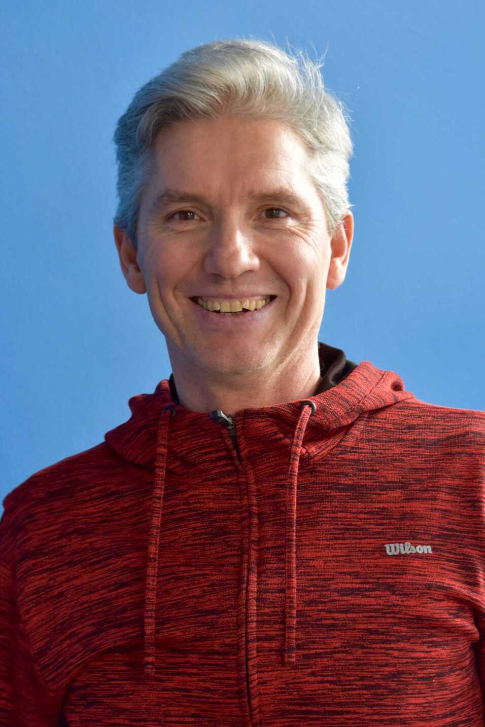 Lächelnder Mann mit grauen Haaren in rotem Sweatshirt vor einem blauen Hintergrund.