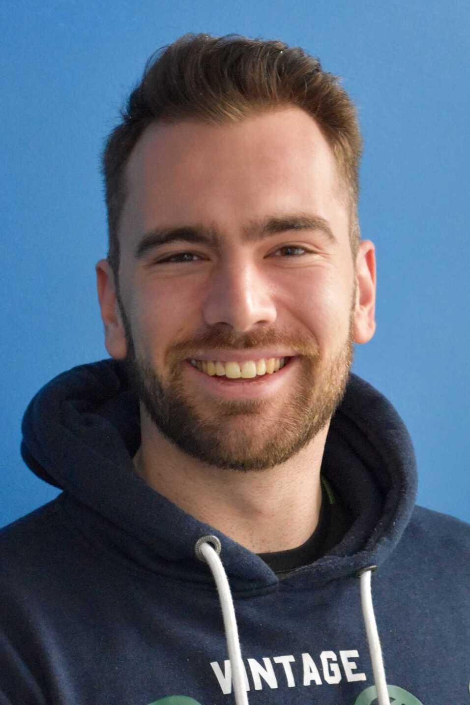 Lächelnder Mann mit kurzem Bart in blauer Kapuzenjacke, vor einer einfarbigen blauen Wand.