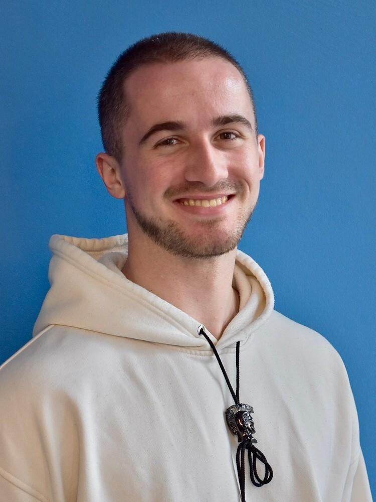 Junger Mann mit Glatze und Bart in einem weißen Hoodie vor einer blauen Wand, lächelt freundlich in die Kamera.