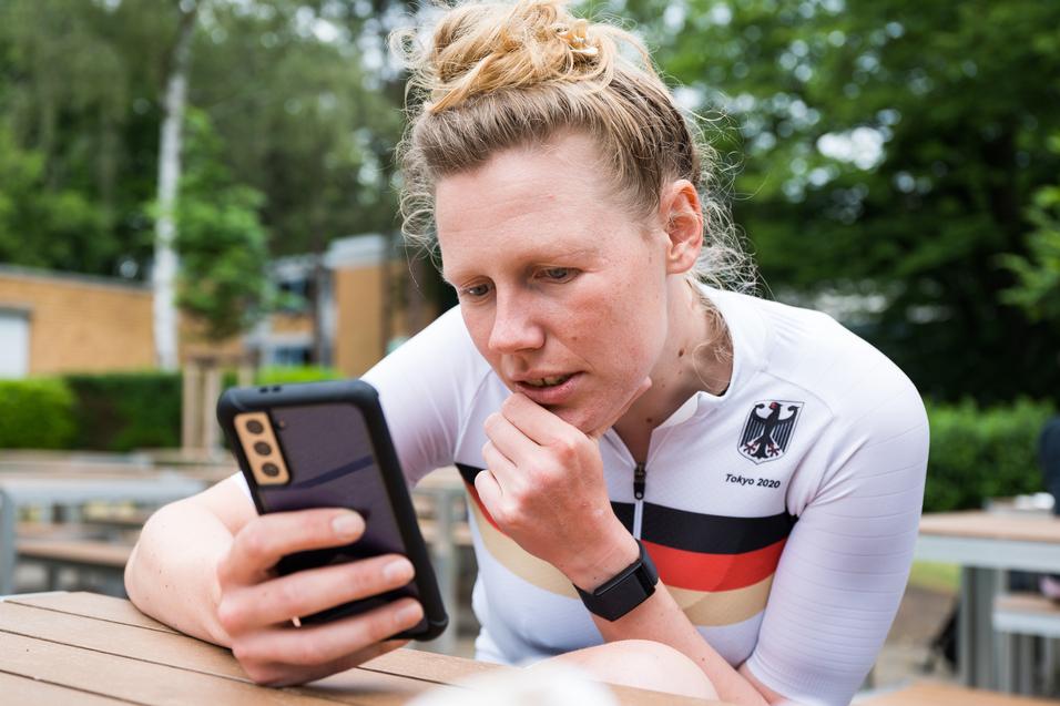 Athletin im weißen Trikot mit schwarzem und rotem Streifen schaut konzentriert auf ihr Smartphone in einer Außenanlage.