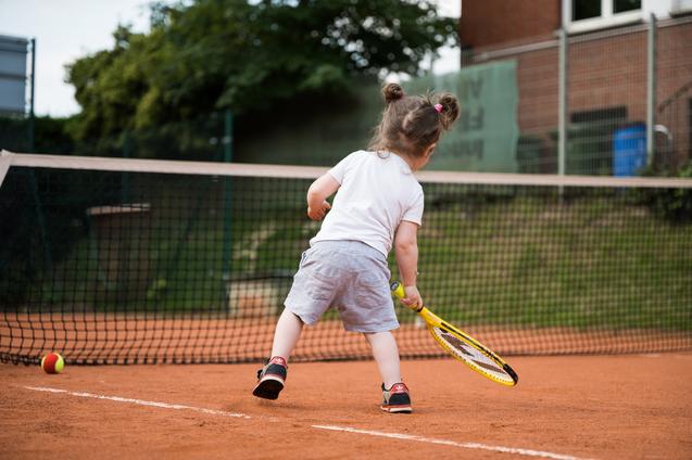 Kind mit Zöpfen schlägt Tennisball auf einem Sandplatz, während es sich auf einen Schlag vorbereitet.